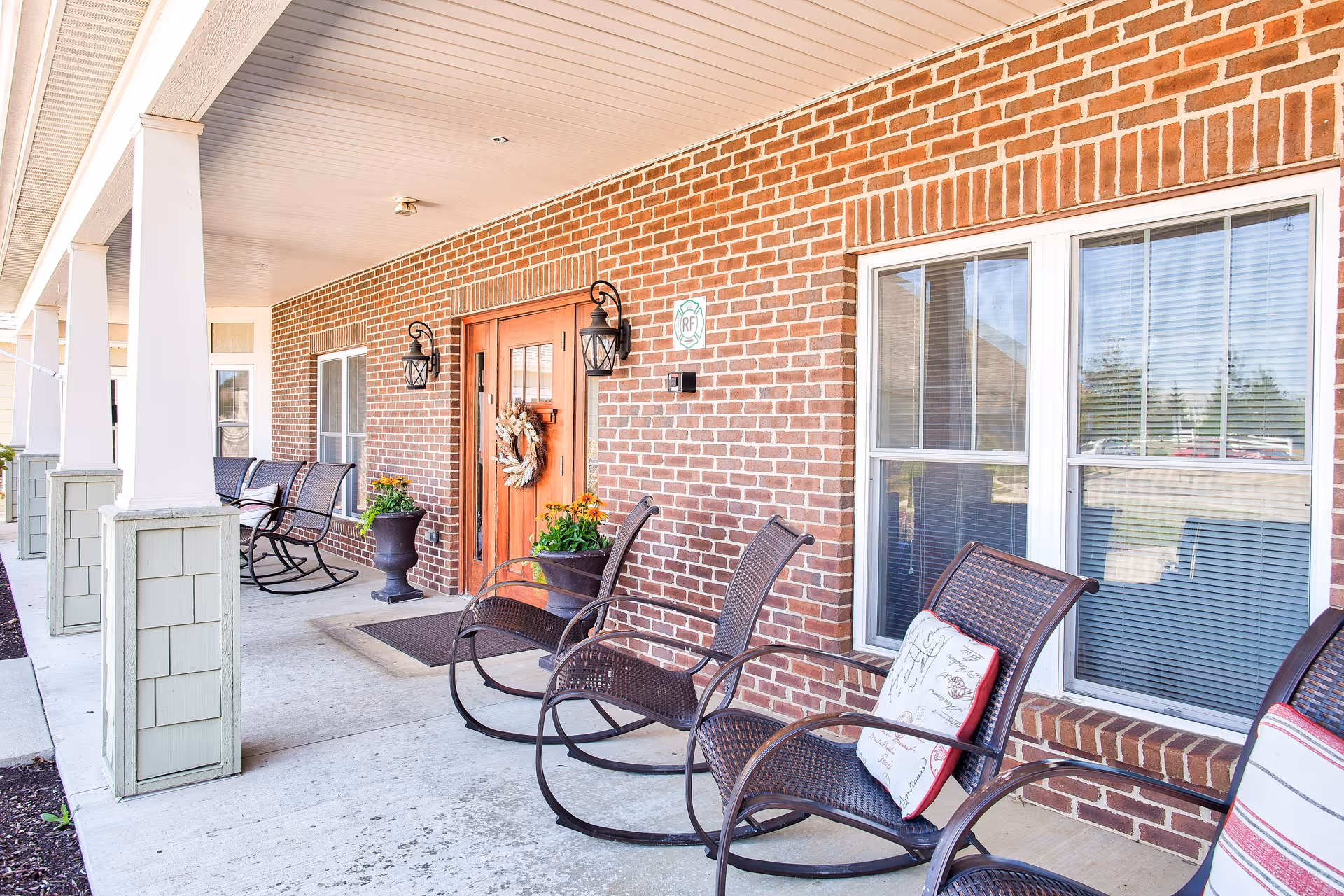 Covered outdoor porch area with brick walls, a wooden door decorated with a wreath, several black rocking chairs with cushions, potted plants, and windows with blinds.