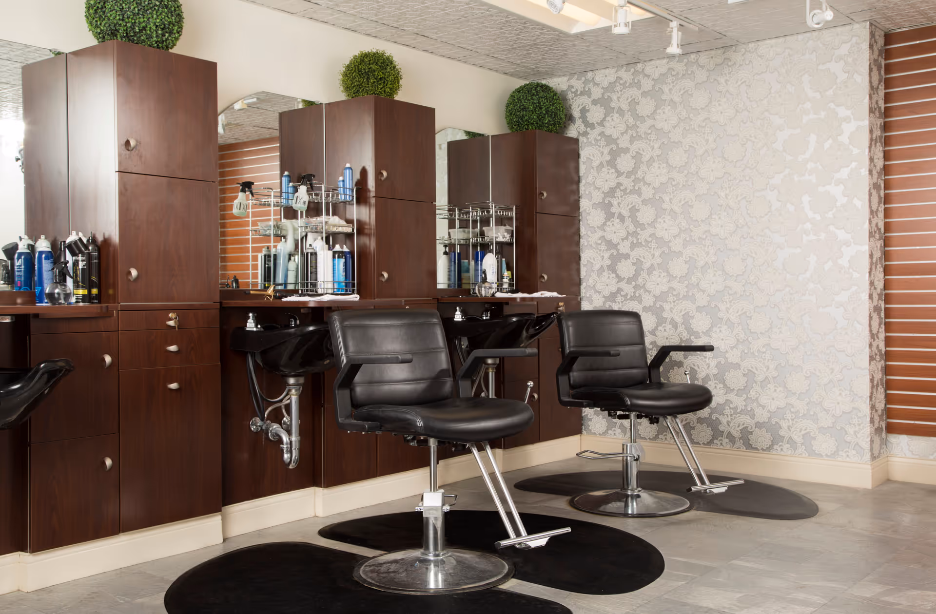Interior view of a salon area with two black salon chairs in front of wooden cabinets and mirrors. The cabinets hold various hair care products, and there are two green decorative plants on top. The wall has a patterned wallpaper and a window with wooden blinds.