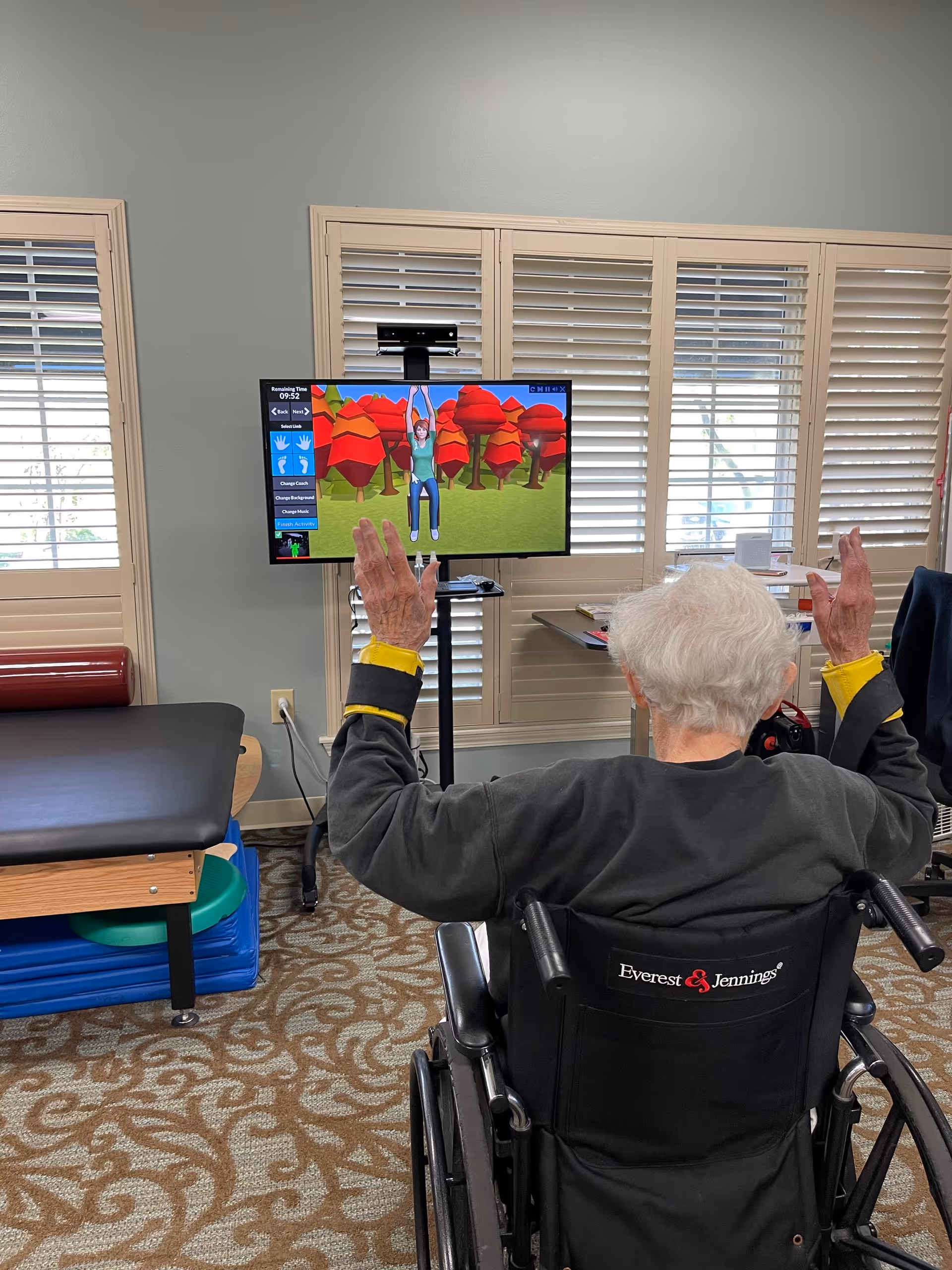 An elderly person with white hair in a wheelchair is raising both hands while facing a TV screen displaying a colorful animated exercise program with a character hanging from a bar in a forest setting. The room has beige walls, windows with white shutters, a therapy table, and exercise equipment.