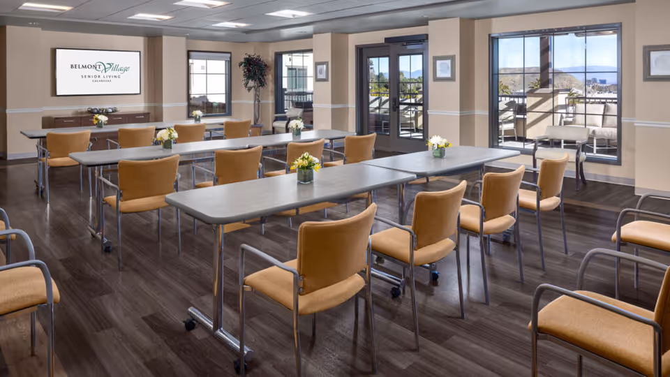 A bright and spacious meeting or activity room with multiple tables arranged in rows, surrounded by orange cushioned chairs on wheels. The room has large windows and glass doors letting in natural light, with a view of an outdoor patio area. There are small flower arrangements on the tables and a large screen on the wall displaying the Belmont Village Senior Living Calabasas logo.