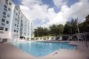 Outdoor swimming pool area with lounge chairs and a white fence, adjacent to a multi-story white building under a partly cloudy sky.