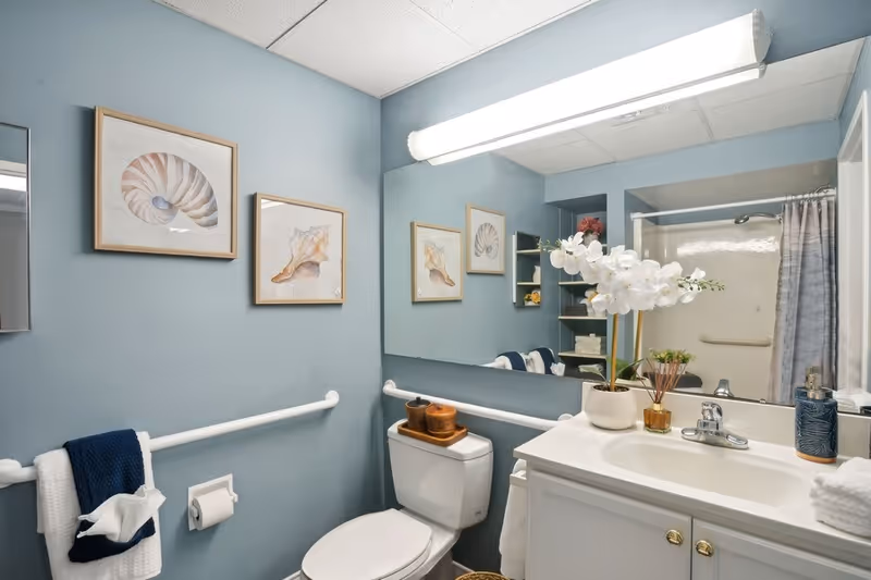 A clean bathroom with light blue walls featuring two framed seashell pictures. There is a white toilet with a wooden tray holding containers on top, a white sink with a faucet, and a large mirror above the sink. A white orchid plant and a reed diffuser are placed on the sink counter. A towel rack with white and navy blue towels is mounted on the wall, and a shower with a curtain is visible in the background.