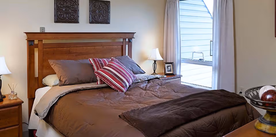 A cozy bedroom with a wooden headboard bed made with brown bedding and pillows, two bedside tables each with a lamp, a window with curtains letting in natural light, and decorative wall art above the bed.