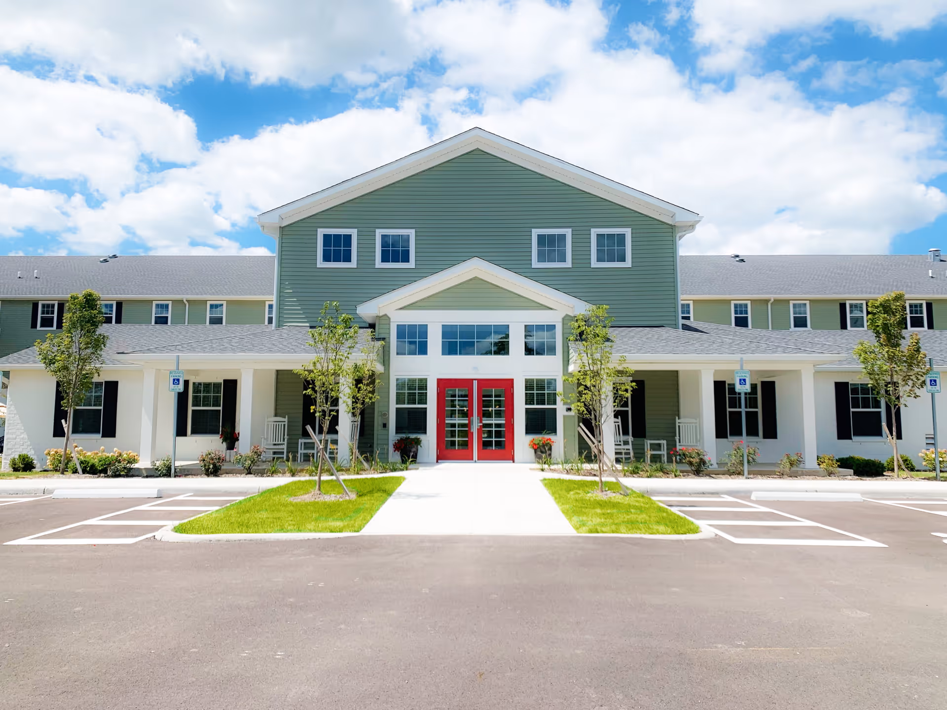 Front exterior of a two-story senior living building with green siding, a covered porch and a red entrance door under a blue sky.
