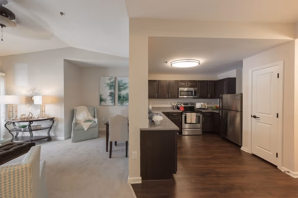 Interior view of a senior living facility showing a combined living and dining area with light-colored walls and carpeted floor. The living area has a light blue armchair with a white throw, a small table with two lamps, and wall art. Adjacent to the living area is a kitchen with dark wood cabinets, stainless steel appliances including a refrigerator, oven, and microwave, and a granite countertop. The kitchen floor is dark wood, contrasting with the carpet in the living area.