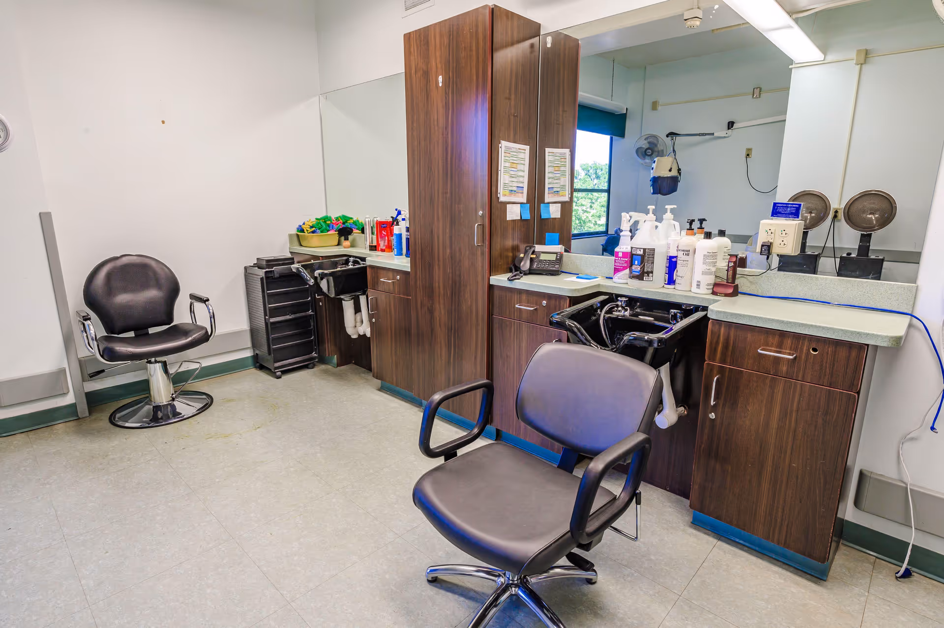 Interior of a hair salon area in a senior living facility with two black salon chairs, a large mirror, wooden cabinets, a sink for washing hair, and various hair care products on the counter.