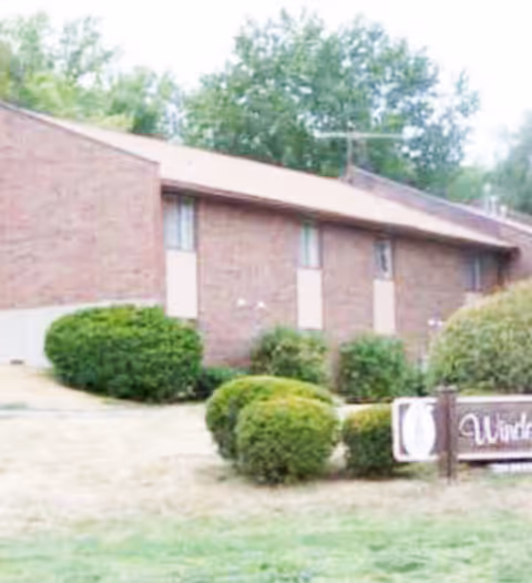 Exterior view of a single-story brick building with several windows, surrounded by green bushes and trees. A sign in front reads 'Windemere Residential Care'.