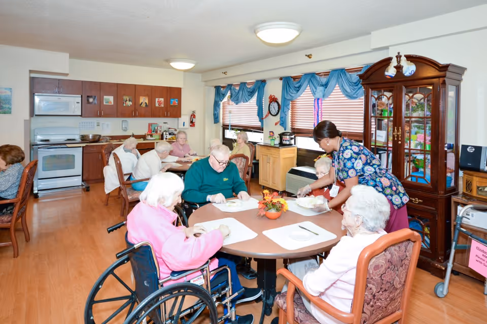 A group of elderly people seated around tables in a dining area of a senior living facility. A caregiver is serving food to one of the residents. The room has wooden flooring, a kitchen area with cabinets and appliances, large windows with blue curtains, and a wooden display cabinet.