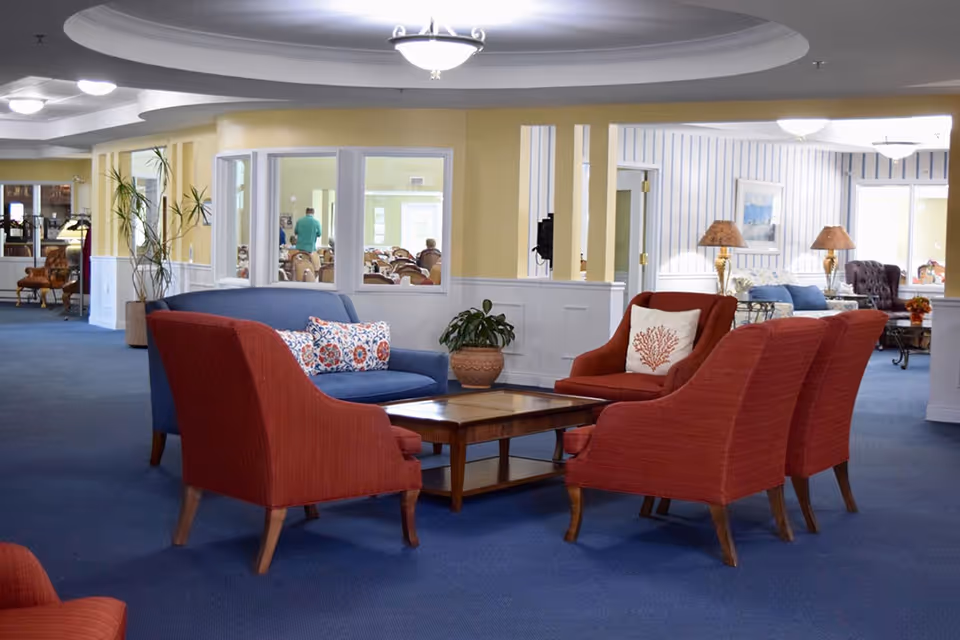 A cozy assisted living facility common area with a blue sofa and four red armchairs arranged around a wooden coffee table. The room has blue carpet, yellow and white walls, and ceiling lights. In the background, there are more seating areas and a dining room visible through windows.