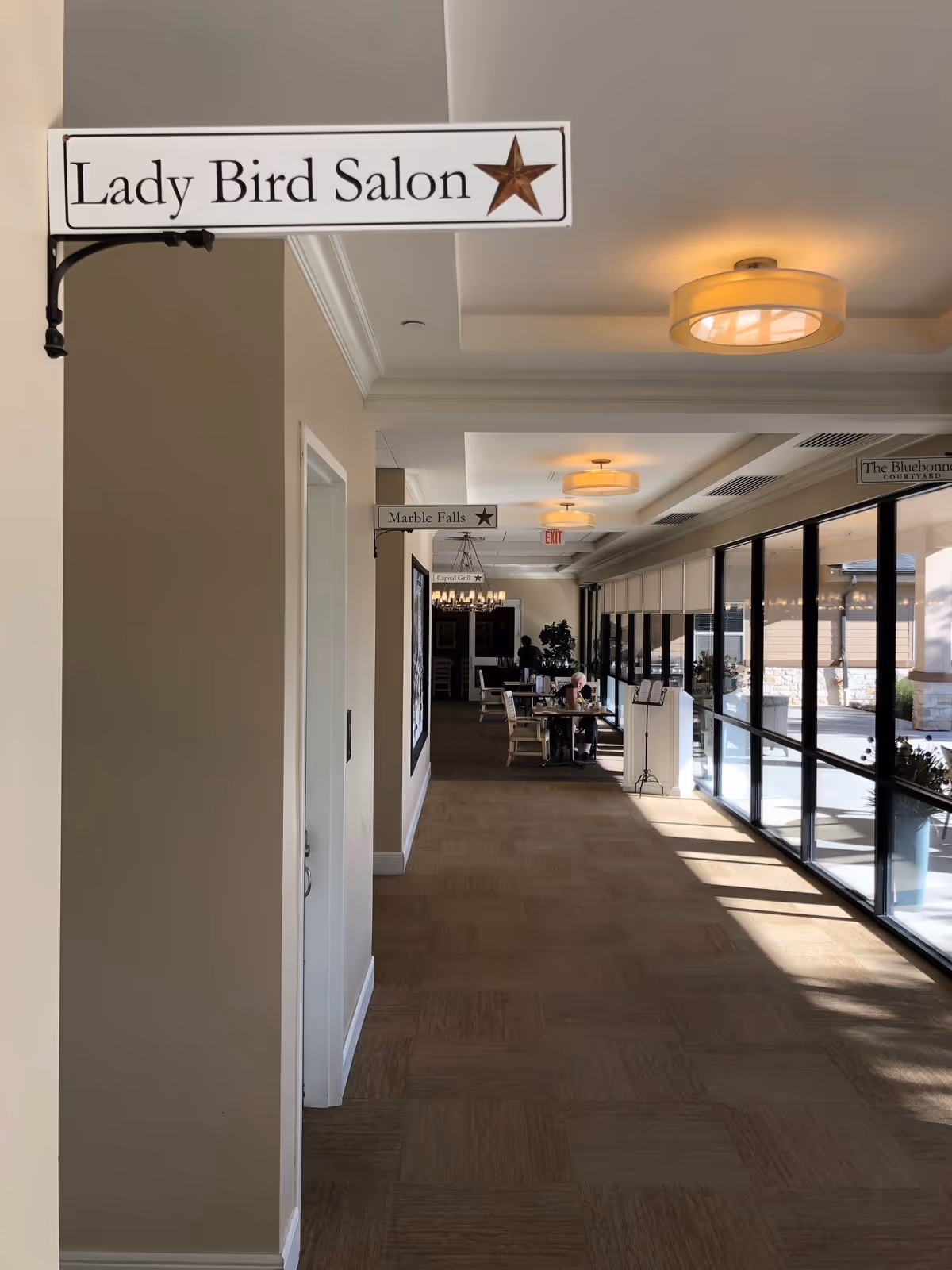 A hallway inside The Legacy at Crystal Falls with signs indicating different rooms including Lady Bird Salon, Marble Falls, and The Bluebonnet. The hallway has large windows on the right side letting in natural light, and there are tables and chairs along the corridor with a few people seated. Ceiling lights illuminate the space.