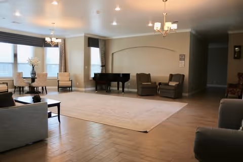 A spacious living room area with wooden flooring, a large beige rug, a black grand piano against the wall, two armchairs, a small table with chairs near windows with curtains, and ceiling light fixtures.