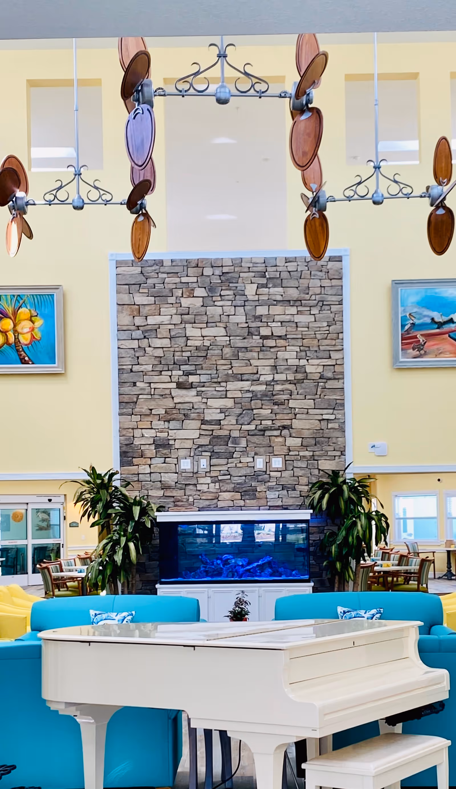 Bright and spacious assisted living common area featuring a white grand piano in the foreground, turquoise sofas with patterned pillows, a large stone accent wall with a built-in aquarium, two large green plants on either side of the aquarium, and colorful paintings on yellow walls. Decorative ceiling fans with wooden blades hang from the ceiling.