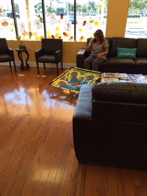 A senior living common room with leather sofas and chairs, a woman seated on a sofa, a colorful activity mat on the hardwood floor and fall-themed window decorations.