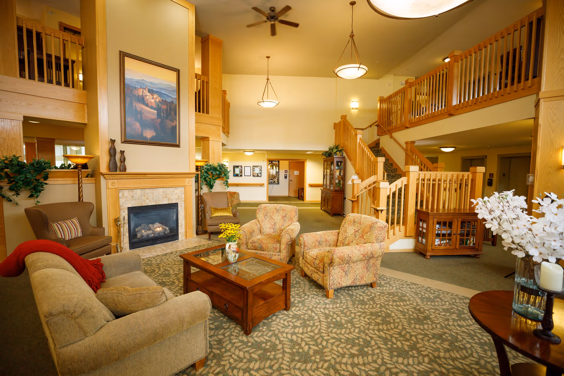 A spacious and warmly decorated senior living facility common area with comfortable upholstered chairs and sofas arranged around a wooden coffee table. There is a fireplace with a framed painting above it, wooden railings on the upper level, a staircase, and soft lighting from ceiling fixtures. The room has a patterned carpet and some decorative plants and flowers.