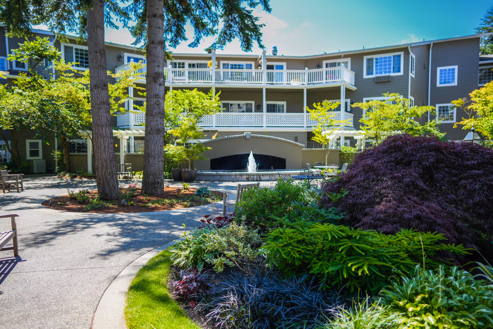 Outdoor courtyard area of a senior living facility with a variety of green plants, trees, and shrubs. There is a paved walkway, benches, and a water fountain in front of a multi-story building with balconies and large windows under a clear blue sky.