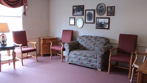 A cozy sitting area with a floral patterned loveseat and three wooden armchairs with red cushions arranged around a small wooden side table with a lamp. The walls are decorated with a collection of framed black and white photographs and artwork. The room has a purple carpet and a window with a maroon valance.
