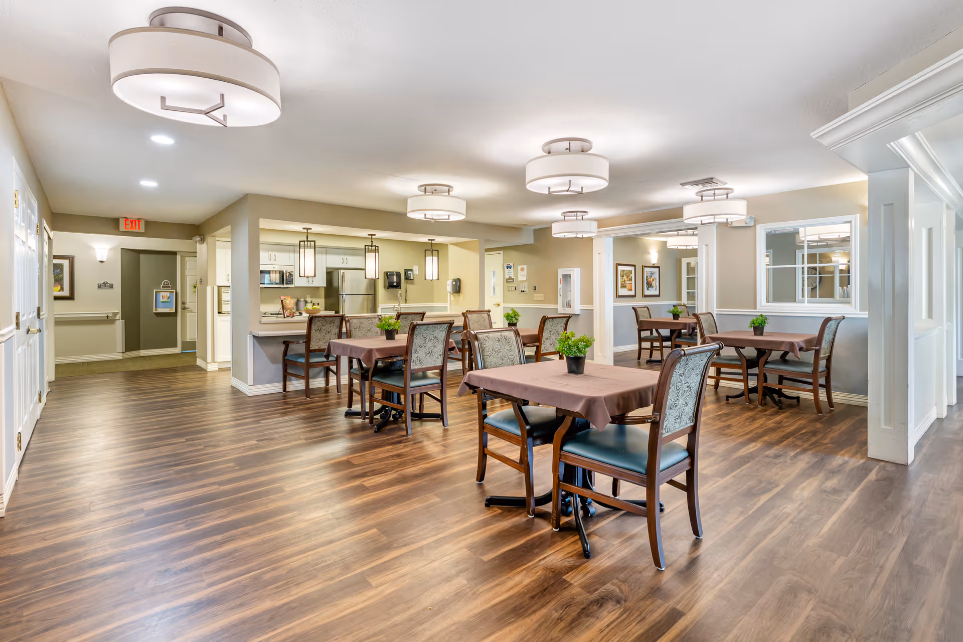 A bright and spacious dining area in a senior living facility with several tables covered with brown tablecloths, each surrounded by wooden chairs with patterned upholstery. The room features wood flooring, modern ceiling lights, and a kitchen area with stainless steel appliances and pendant lights in the background. There are framed pictures on the walls and a large mirror reflecting part of the room.