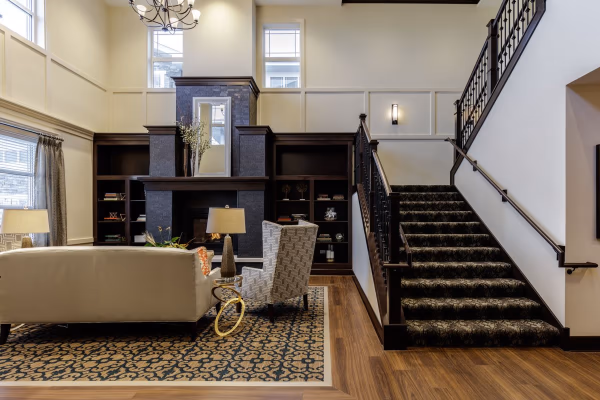 A spacious living room area with a high ceiling, featuring a large dark fireplace with built-in shelves on either side. In front of the fireplace, there is a white sofa and a patterned armchair around a small round table with a lamp. A patterned rug covers part of the wooden floor. To the right, a carpeted staircase with dark wooden railings leads upstairs. Large windows allow natural light to fill the room.