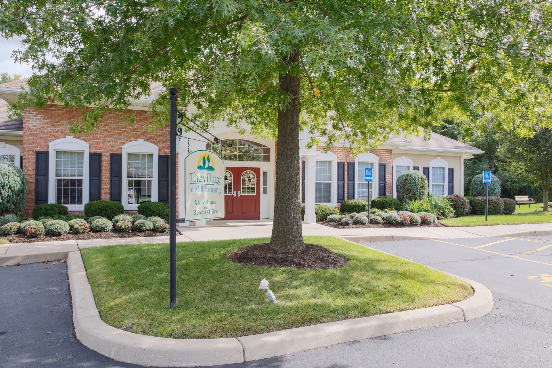 Front exterior of The Village Townhomes clubhouse with a hanging sign, large tree, landscaped beds, and handicap parking spaces.