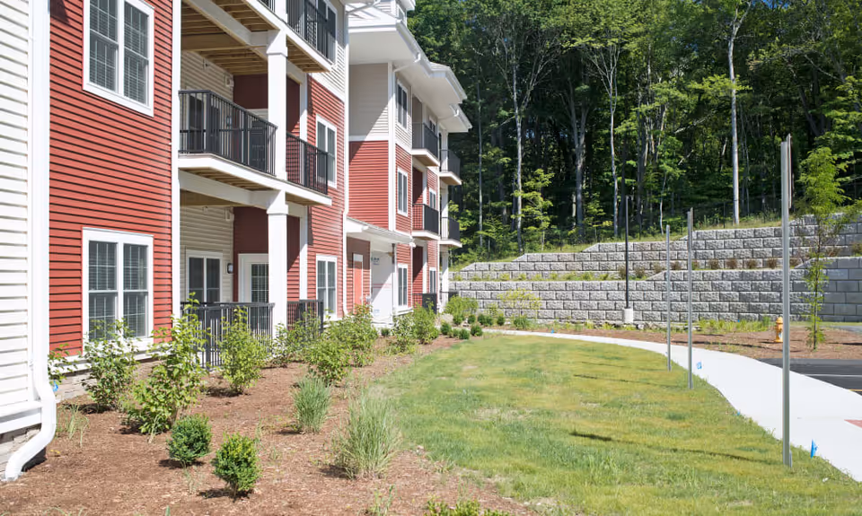 Exterior view of a multi-story residential building with red and white siding, balconies with black railings, and a landscaped area with small bushes and grass. A sidewalk runs alongside the building, and there is a retaining wall with trees in the background.