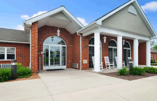 Front exterior view of a brick building with white columns and a covered entrance. There are rocking chairs on the porch and a clear blue sky above.