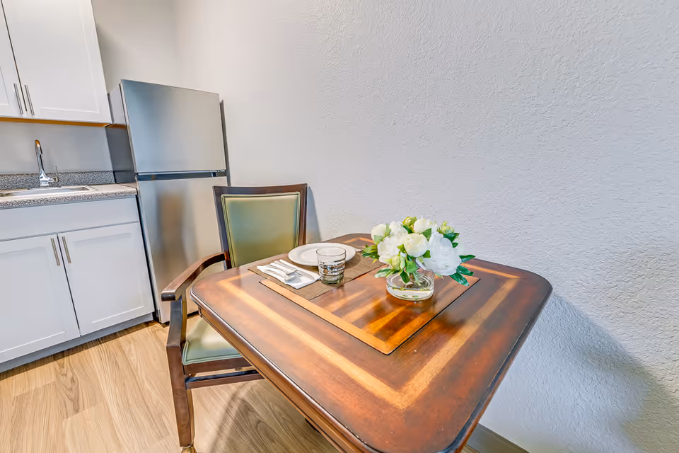 Small dining nook with a wooden table set for one and a vase of white flowers beside a compact kitchenette with a stainless refrigerator and white cabinets.