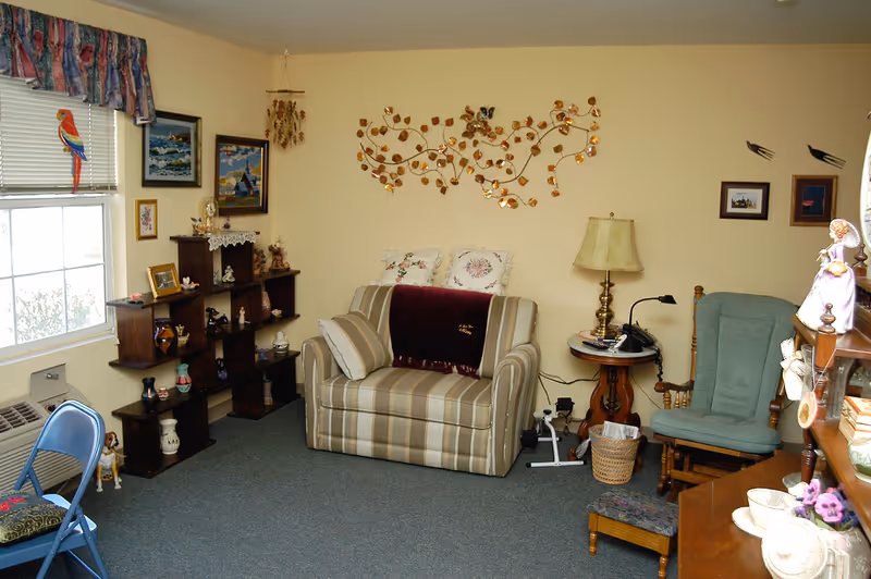 Cozy living room with a striped loveseat, decorative wall art, shelving with knickknacks, a window, and assorted chairs and tables.