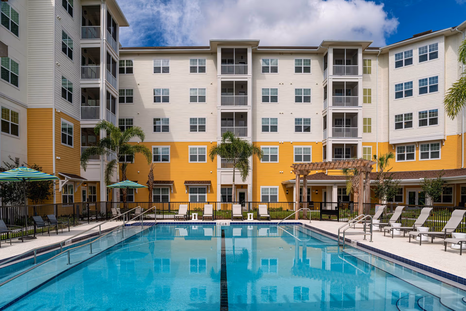 Outdoor swimming pool with clear blue water surrounded by lounge chairs and umbrellas, set in front of a multi-story residential building with white and yellow exterior walls and balconies under a partly cloudy sky.