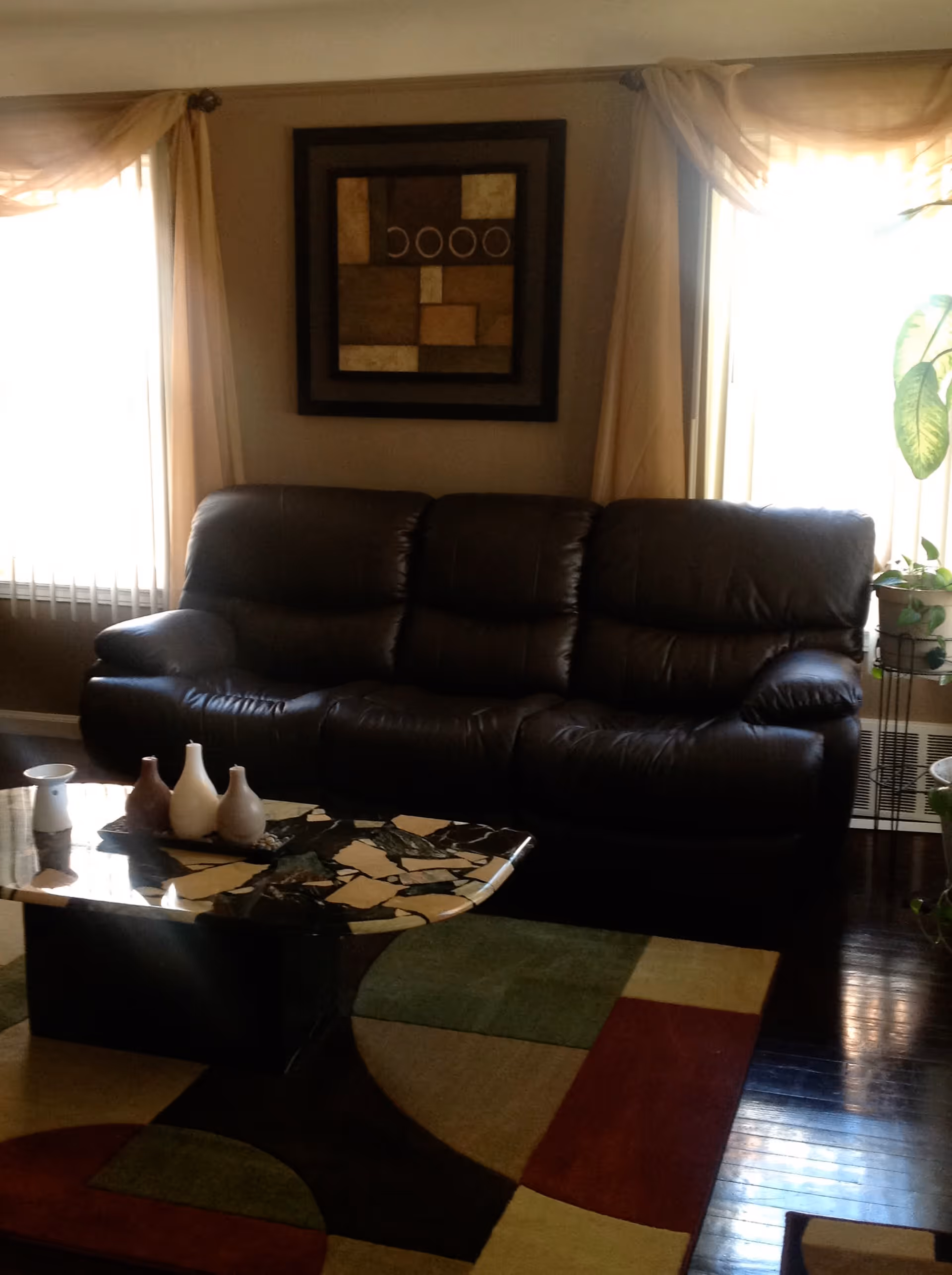 A living room with a dark brown leather sofa positioned between two windows with beige curtains. Above the sofa hangs a framed abstract artwork with geometric shapes. In front of the sofa is a coffee table with a mosaic-style top and three decorative vases. The floor features a multicolored area rug with green, red, beige, and brown sections. A potted plant is visible to the right near the window.