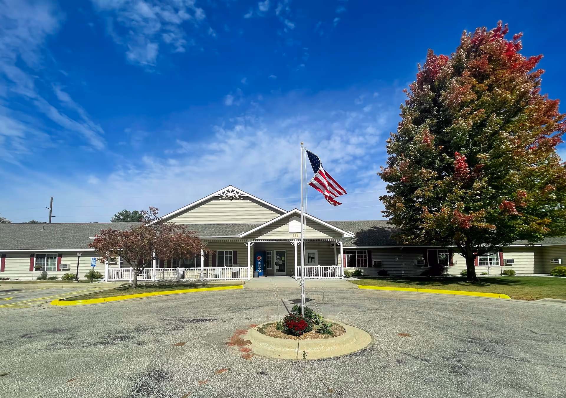 Front exterior view of Park Lane Estates building with a circular driveway, an American flag on a flagpole in the center, and trees with autumn foliage on either side under a partly cloudy blue sky.