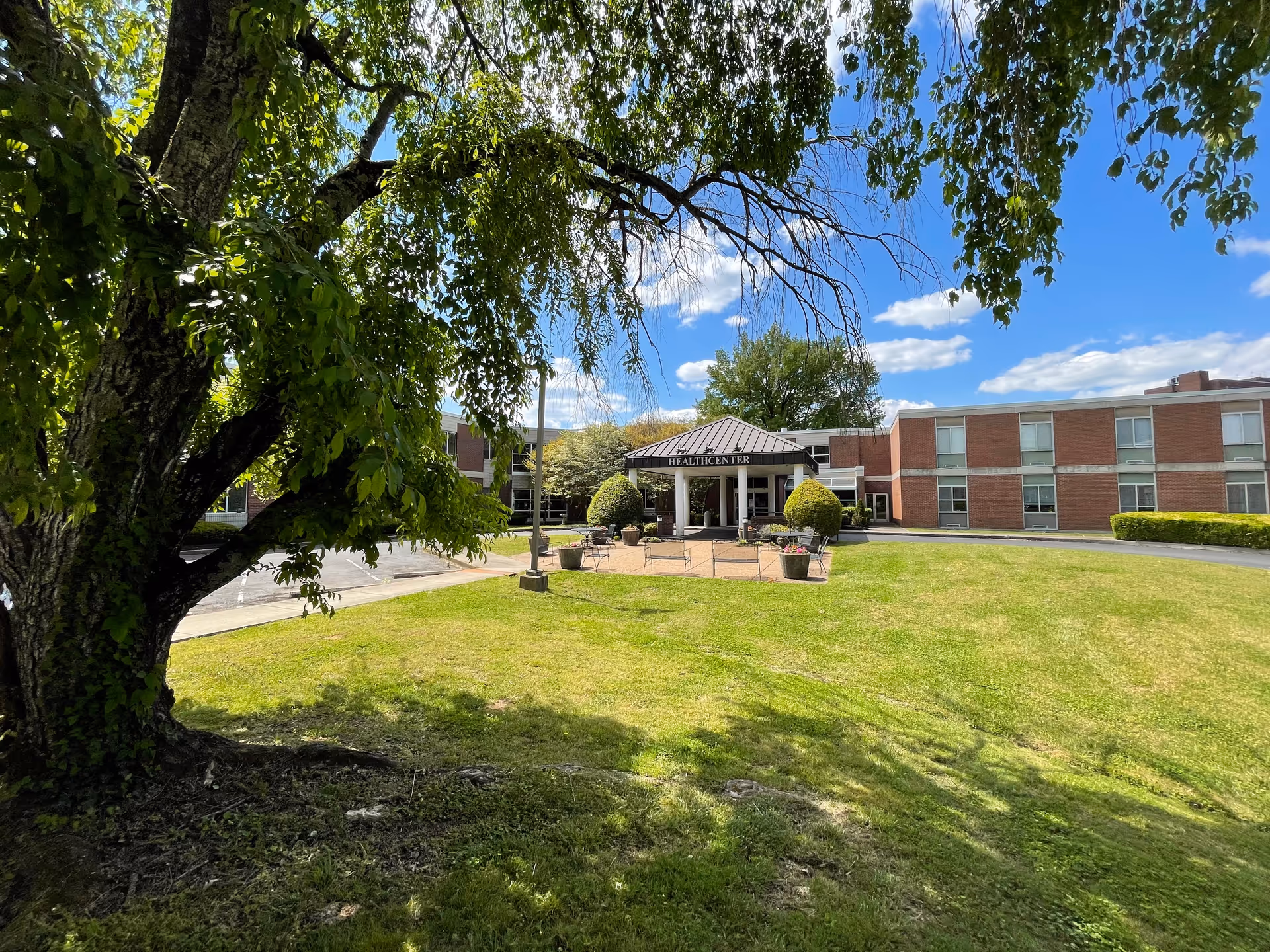 View of a health center building with a covered entrance labeled 'HEALTHCENTER' surrounded by a well-maintained lawn and trees under a blue sky with scattered clouds.
