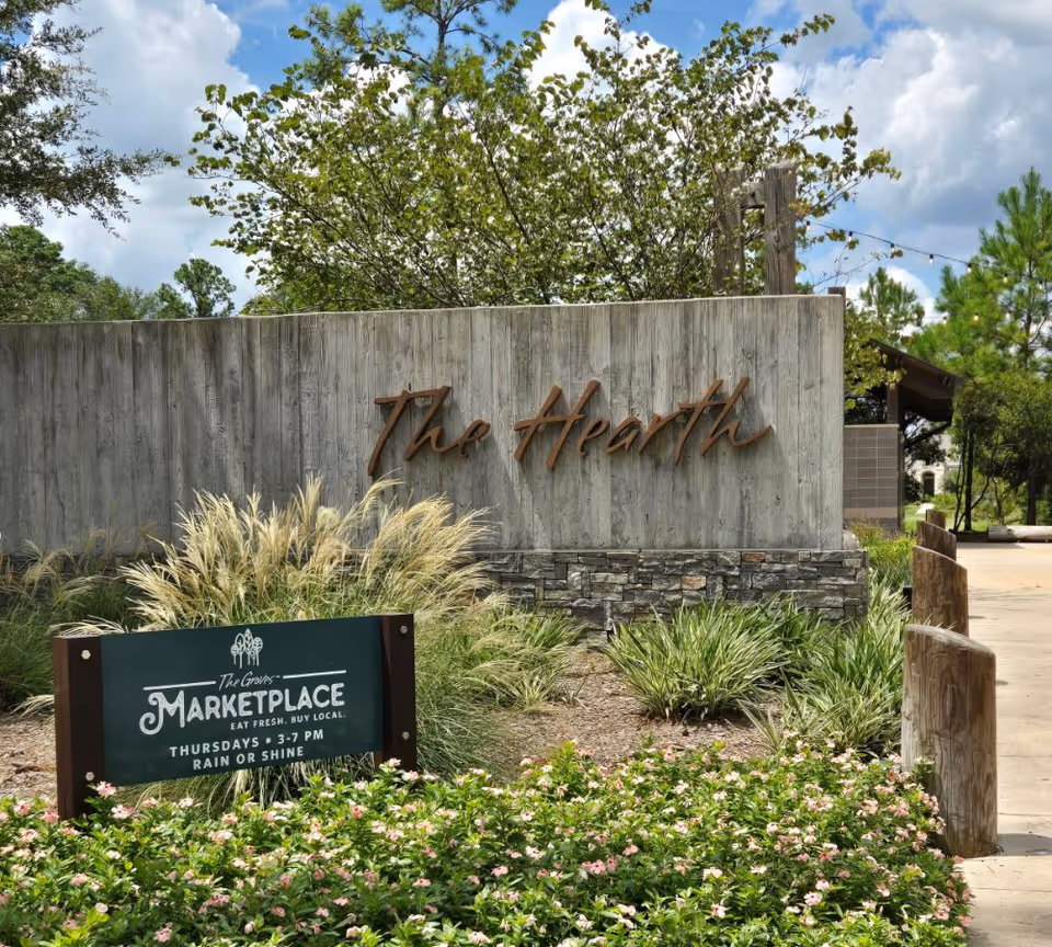 An outdoor entrance wall reading "The Hearth" with landscaped plants and a smaller "Marketplace" sign in front under a partly cloudy sky.