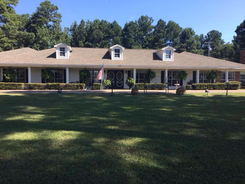 Front exterior view of a single-story retirement lodge building with a well-maintained lawn, several windows with black shutters, hanging plants, and an American flag near the entrance. The building is surrounded by tall trees under a clear blue sky.