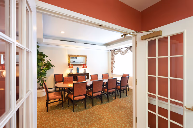 View through open double glass-paneled doors into a dining room with a long rectangular table surrounded by ten upholstered chairs. The room has a patterned carpet, a large window with decorative curtains, a sideboard with two lamps and a mirror above it, and a potted plant in the corner.