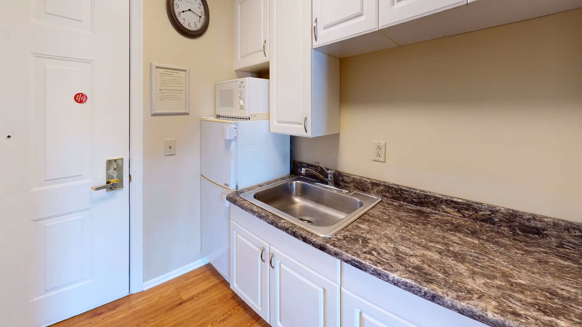 Small kitchen area with a white door on the left, a clock on the wall, a white refrigerator with a microwave on top, white cabinets above and below a brown marble countertop with a stainless steel sink, and wooden flooring.