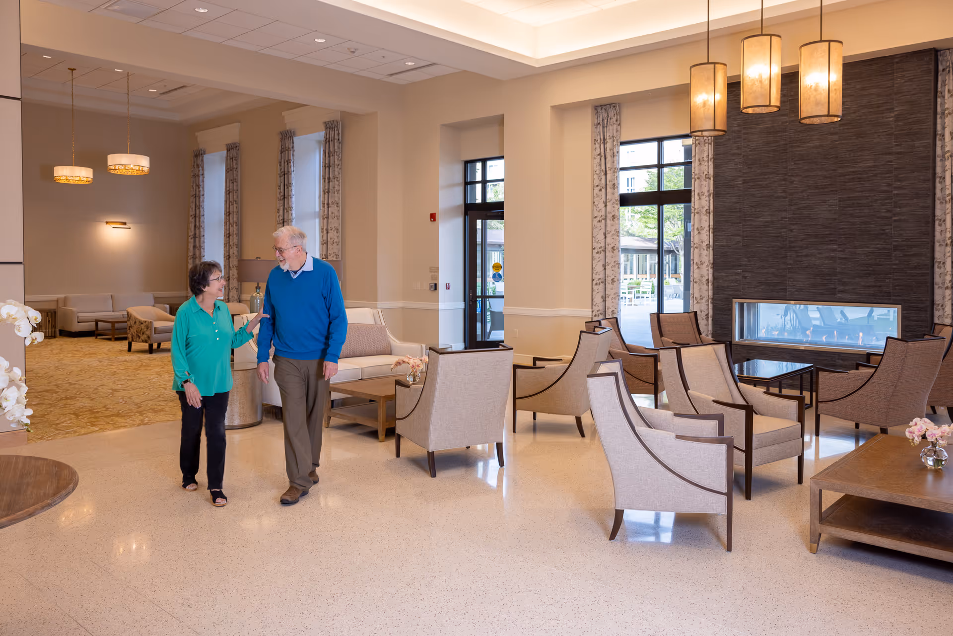 Two elderly people walking and talking in a spacious, well-lit lounge area with beige walls, large windows, comfortable armchairs, sofas, a modern fireplace, and hanging pendant lights.
