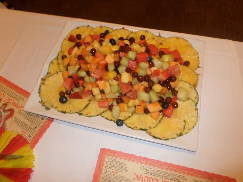 A rectangular white plate with a colorful fruit salad consisting of pineapple slices arranged around the edge and a mix of diced melon, watermelon, and berries in the center, placed on a white tablecloth with red-bordered placemats nearby.