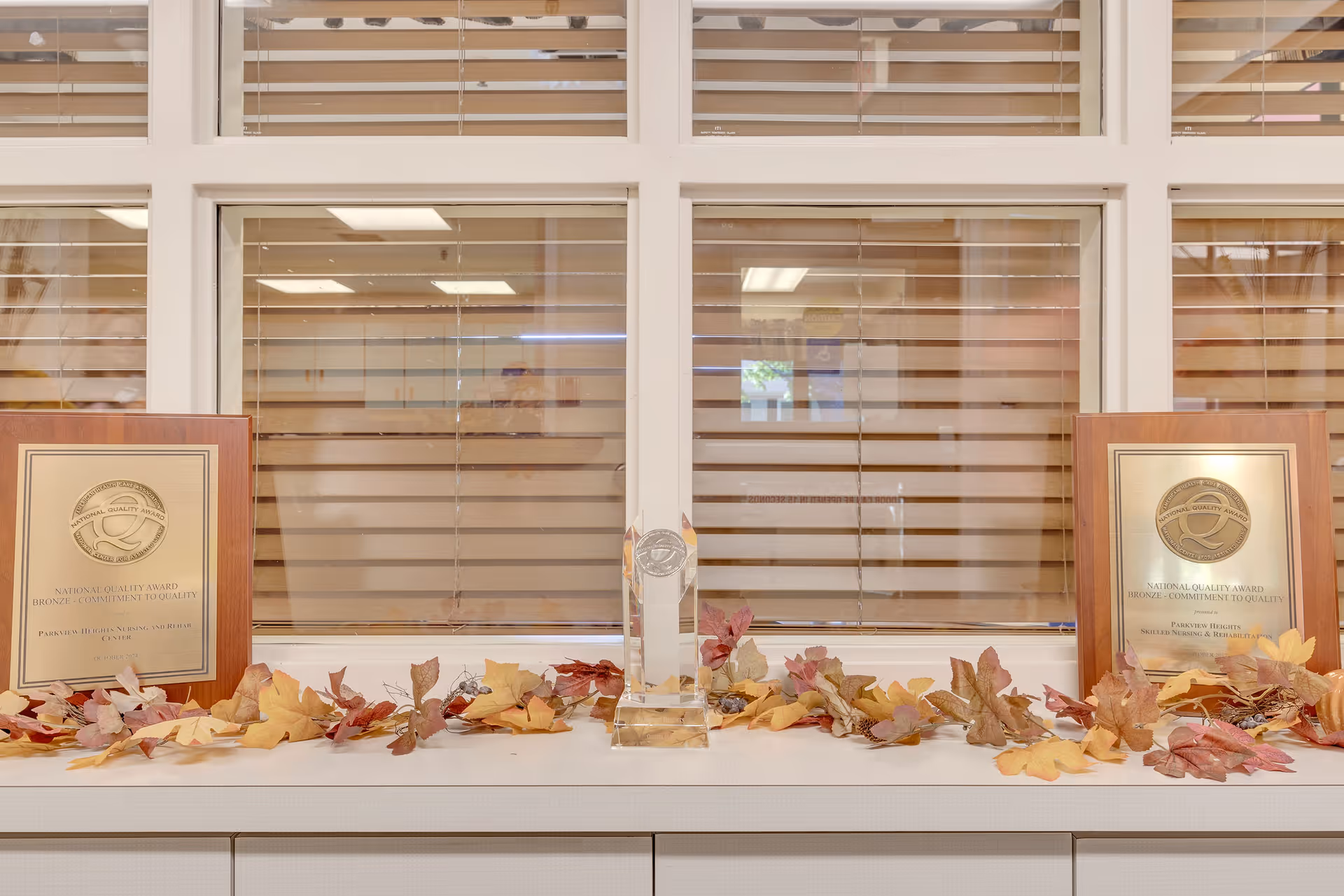 A display shelf with two National Quality Award plaques on either side and a clear glass trophy in the center, decorated with autumn leaves. Behind the shelf is a window with horizontal blinds partially closed, showing a reflection of a room with ceiling lights.
