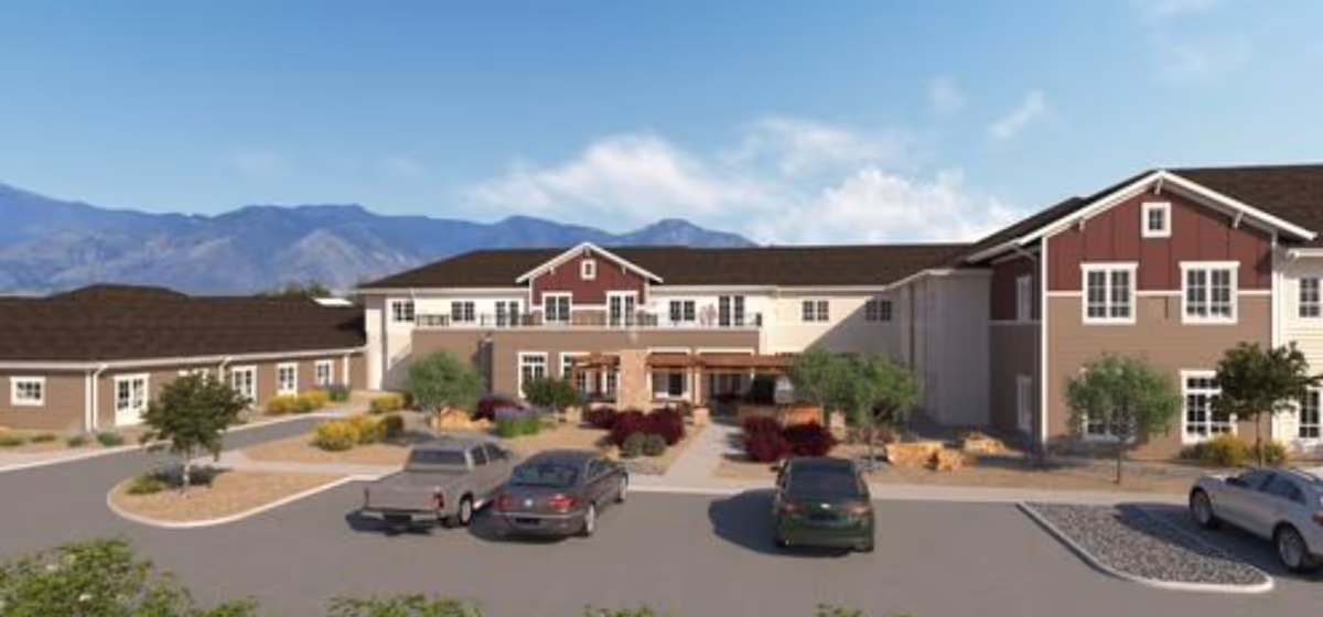 Front exterior of a two-story senior living facility with a landscaped entrance, parked cars, and mountains in the background.