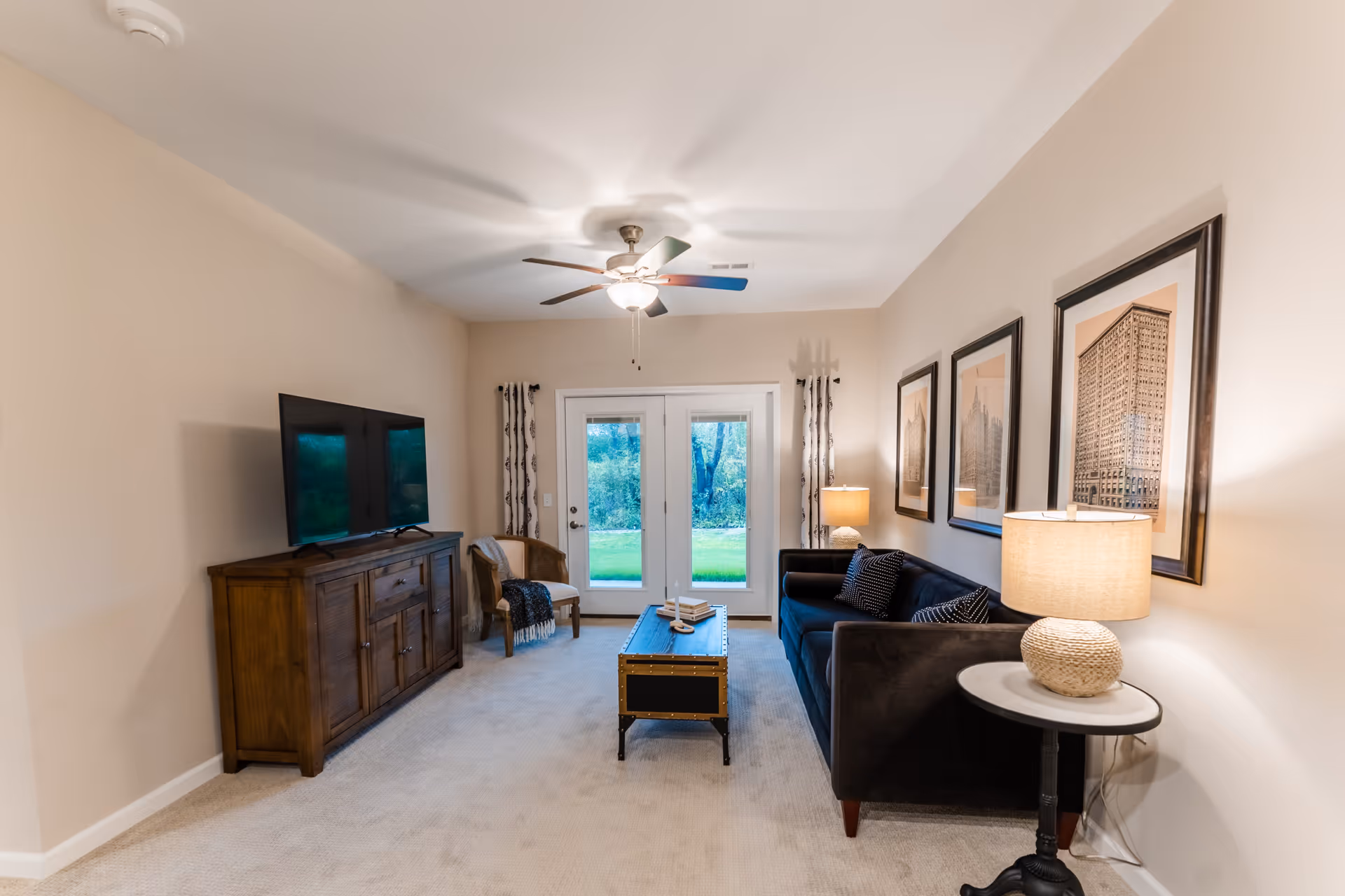 A cozy living room with a dark blue sofa, three framed pictures on the wall, a wooden TV stand with a flat-screen TV, a small round side table with a lamp, a wooden chair with a throw blanket, a ceiling fan with light, and double glass doors leading outside to a green yard.