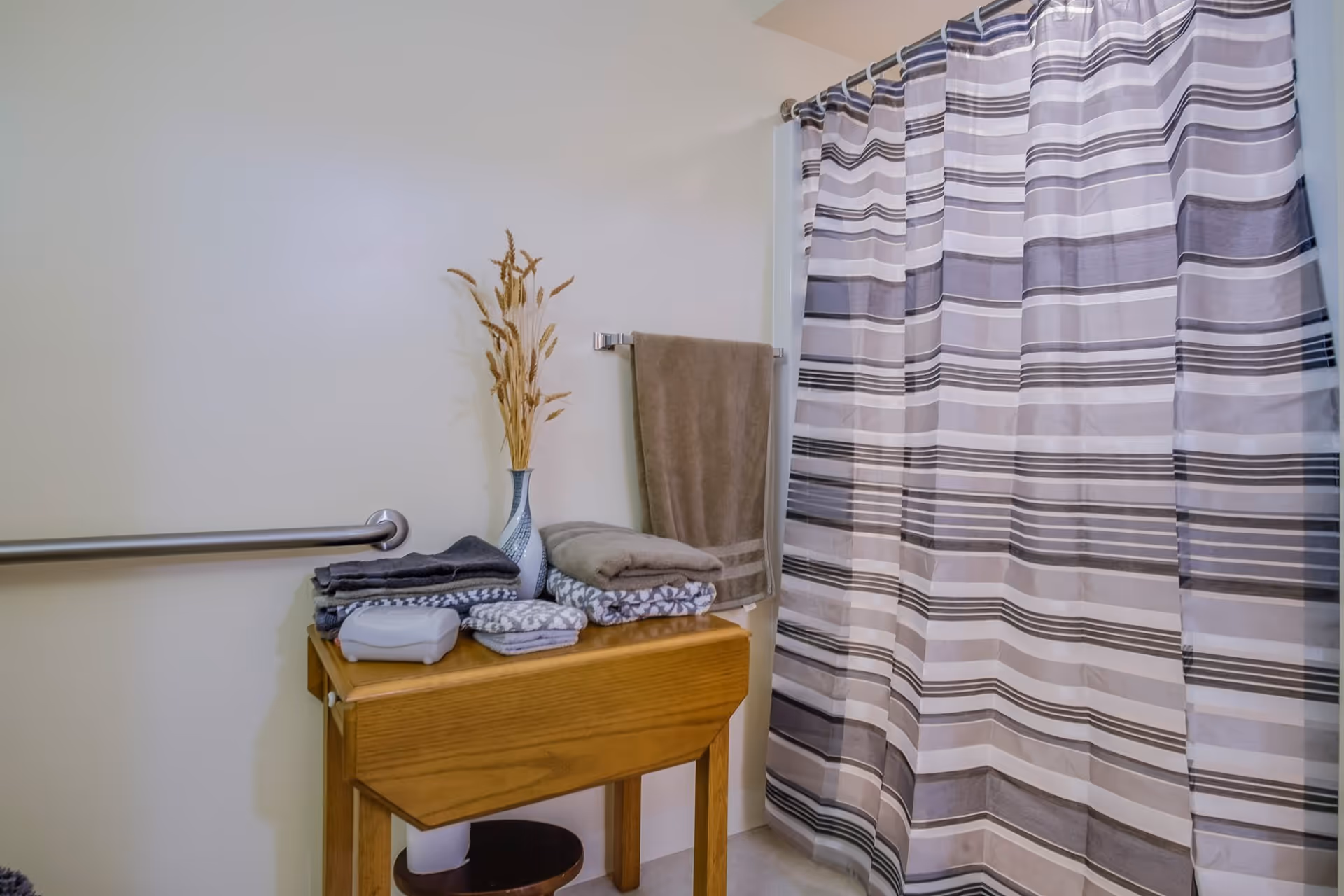 Bathroom interior with a striped shower curtain, a wooden table holding folded towels and a vase with dried plants, a towel hanging on a wall-mounted rack, and a grab bar on the wall.