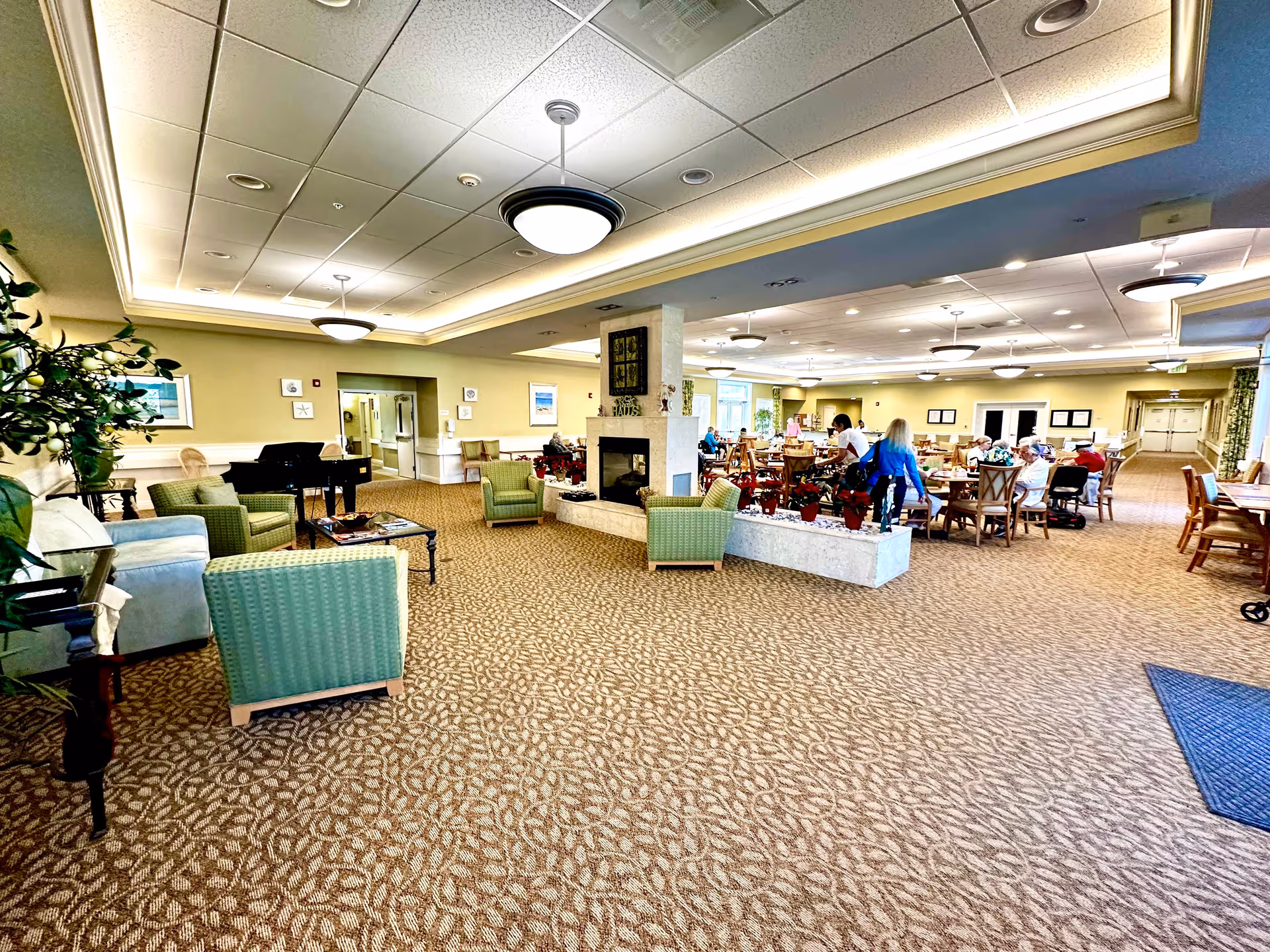 A spacious senior living common area with patterned carpet, green and beige armchairs, a central fireplace with plants around it, and a grand piano in the corner. Several seniors and staff are seated at dining tables in the background, under ceiling lights and a drop ceiling with recessed lighting.