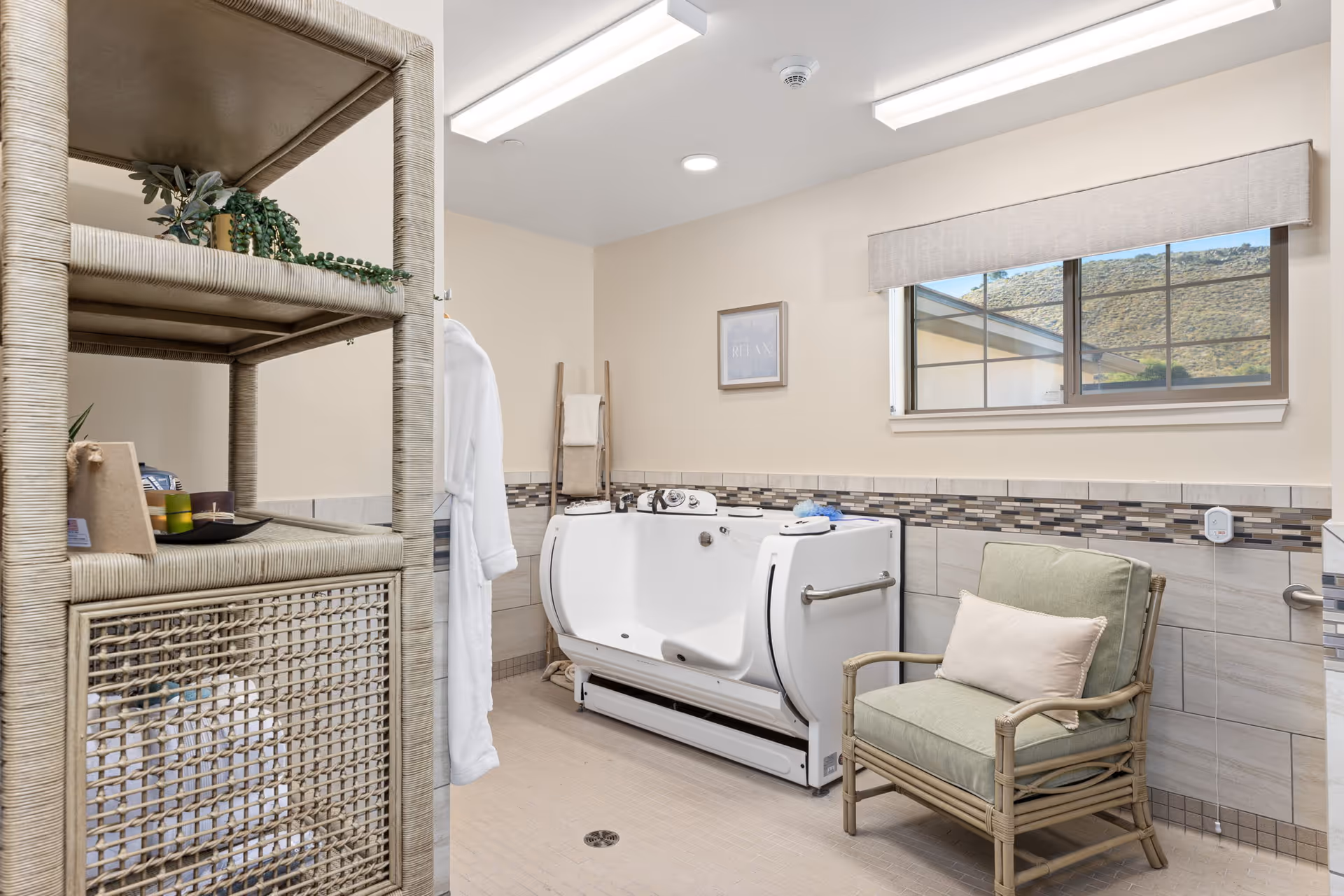 A bright and clean bathroom with a walk-in bathtub, a cushioned armchair with a pillow, a wicker shelving unit with decorative items, a white bathrobe hanging on the wall, and a window showing a view of hills outside.
