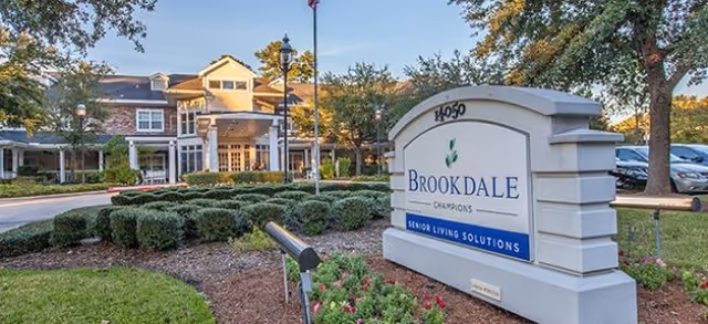 Landscaped entrance of the Brookdale Champions senior living community showing a front sign, driveway, and building facade.