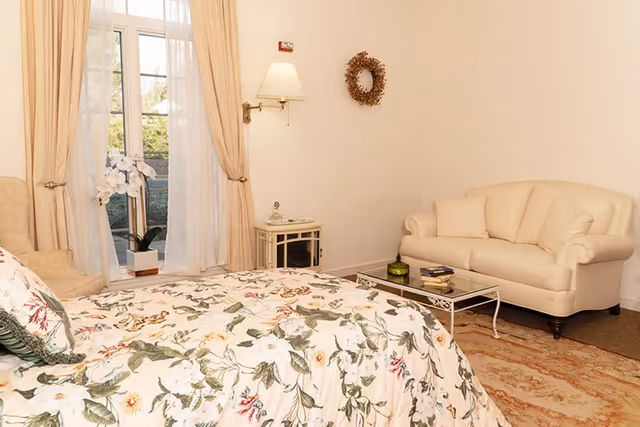 Bright bedroom with a floral bedspread, a beige sofa and glass coffee table, a nightstand and a window with curtains.