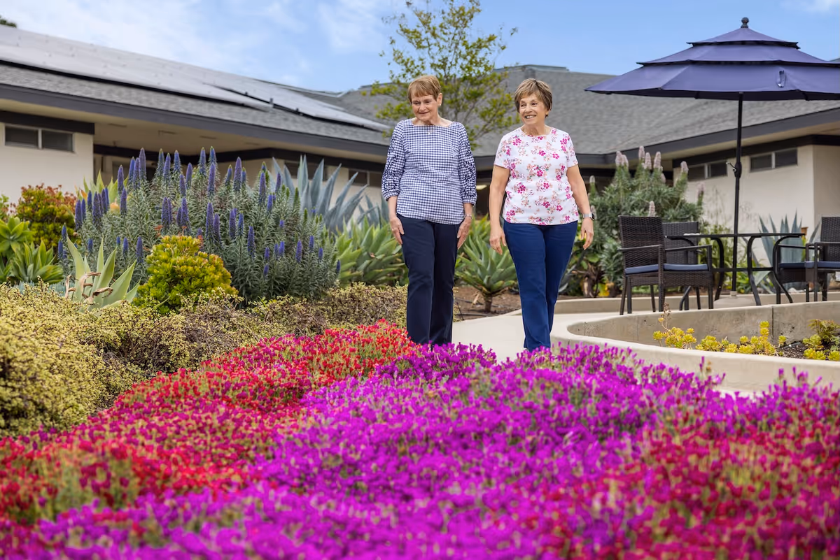 Two women walking on a paved path beside vibrant purple and pink flower beds and patio seating outside a single-story building.