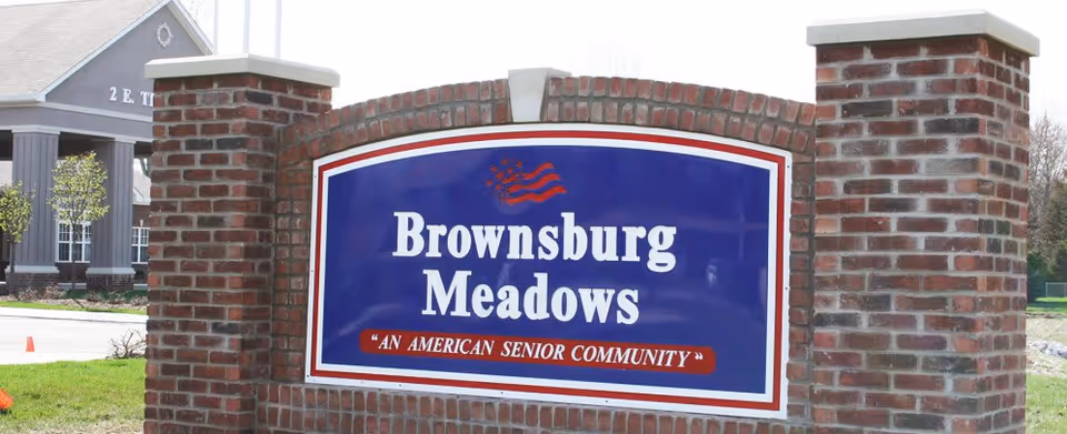 A large brick sign with a blue background and white text that reads 'Brownsburg Meadows' with a smaller red banner below stating 'An American Senior Community'. The sign is situated outdoors with a building and some trees visible in the background.