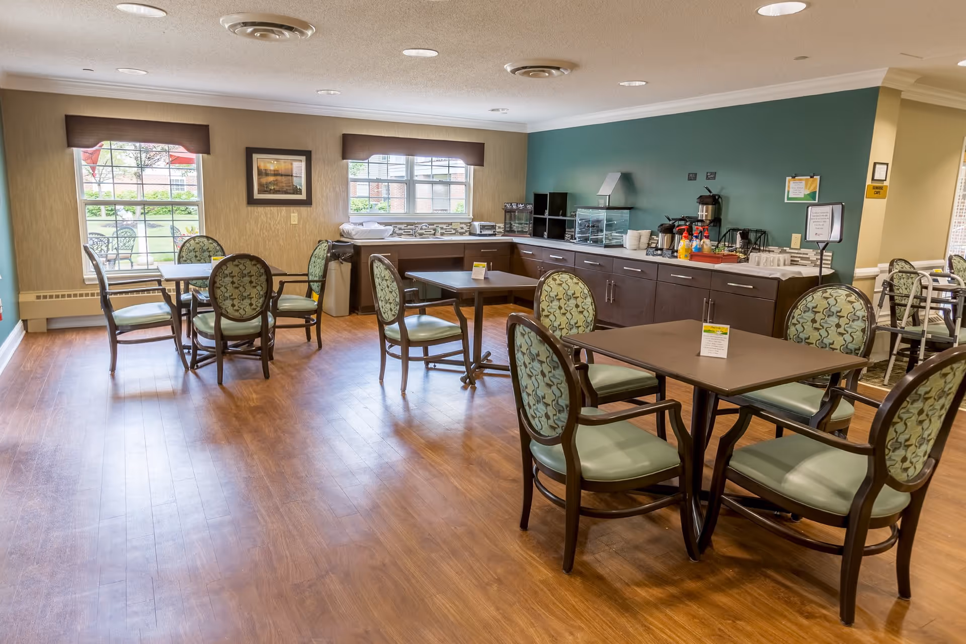 A bright dining area with several tables and chairs arranged on a wooden floor. The room has large windows with brown valances letting in natural light. Against one wall is a counter with various coffee and beverage supplies, including a coffee maker and cups. The walls are painted in a combination of beige and green.