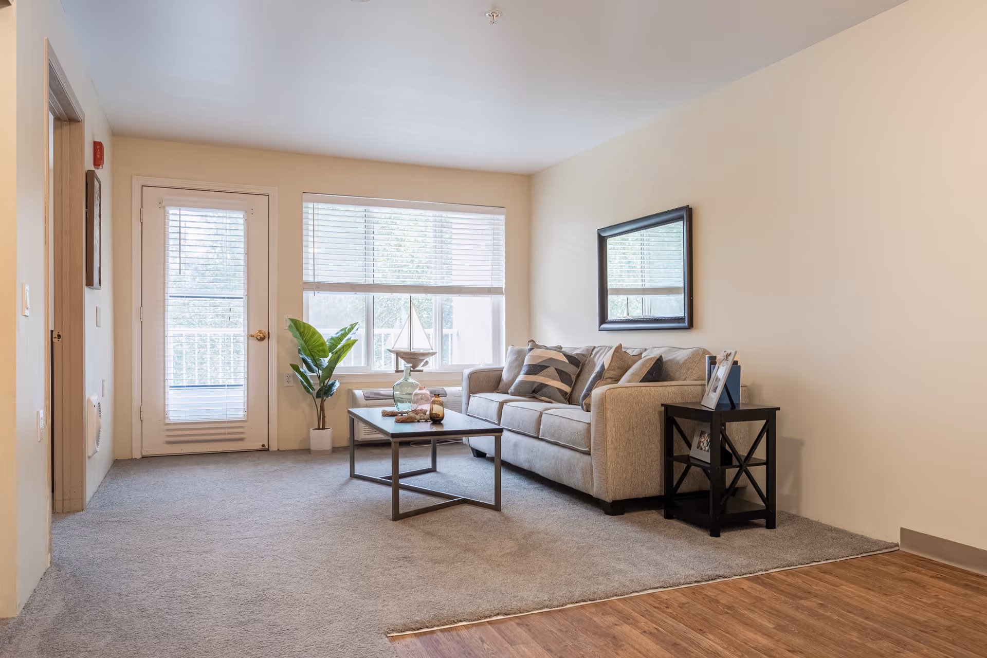A bright living room with a beige sofa adorned with patterned cushions, a black coffee table with decorative items, a small black side table with photo frames, a large window with blinds, a glass door leading outside, and a potted plant near the window.
