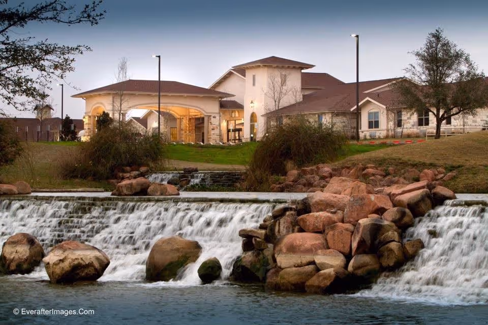 View of a senior living facility building behind a cascading waterfall with large rocks and grassy landscaping, under a clear sky at dusk.